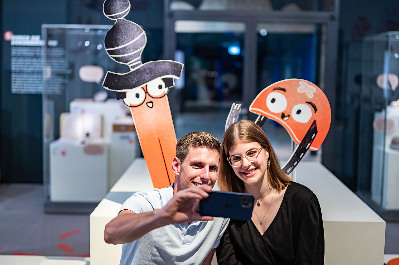 Zwei Besucher*innen machen ein Selfie vor Aufstellern von Helm und Schwert.
